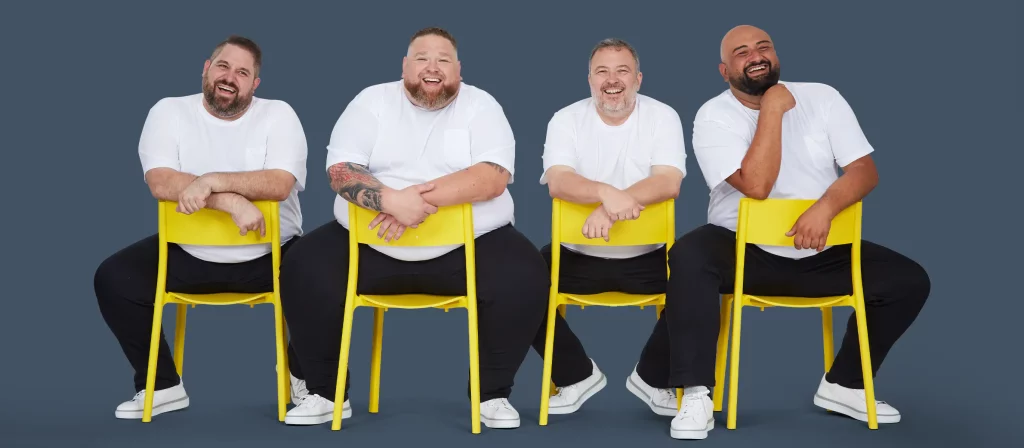 Vier Männer in Übergröße sitzen auf Stühlen, gekleidet in weiße T-Shirts.