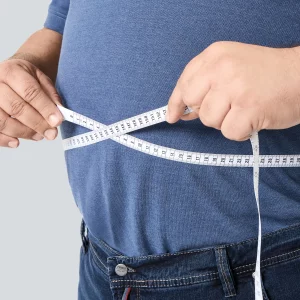 Mann misst seinen Bauchumfang mit einem Maßband, trägt Jeans und ein blaues Poloshirt.