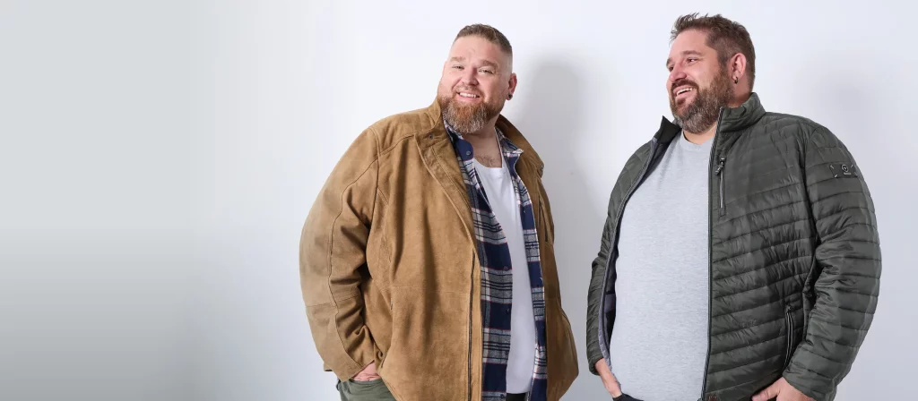 Zwei Männer mit Übergröße tragen stylische Outfits. Der linke in Karohemd und Lederjacke, der rechte in Steppjacke und T-Shirt.