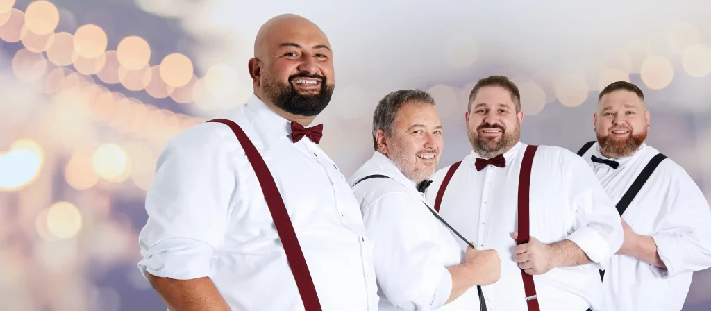 Vier Männer in weißen Hemden, mit Hosenträgern und Fliegen, die in die Kamera lächeln