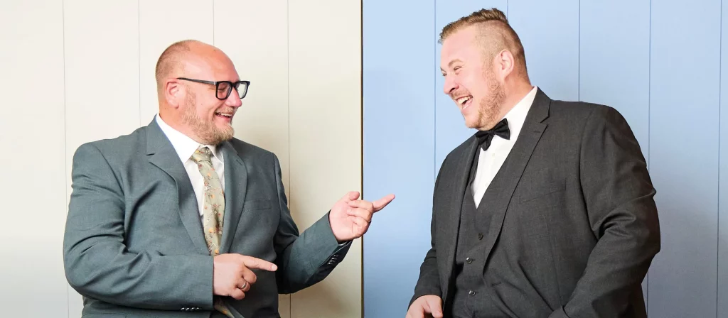 Zwei Männer mit Übergewicht tragen elegante Sakkos – ein grünes Sakko mit Krawatte und ein anthrazitfarbenes Sakko mit Fliege – lachen miteinander.