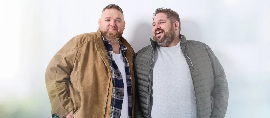 Zwei Männer mit Übergröße: links in Lederjacke mit offenem Karohemd und weißem T-Shirt, rechts in grüner Steppjacke mit grauem Pullover