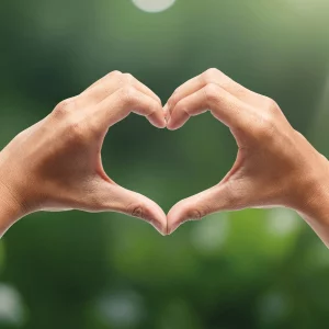 Zwei Hände formen ein Herz vor einem grünen, verschwommenen Hintergrund – Symbol für Nachhaltigkeit und Fairness.