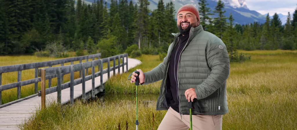 Mann mit Wanderstöcken steht am Fuße eines Berges, mit einer Brücke über einen Bach im Hintergrund.