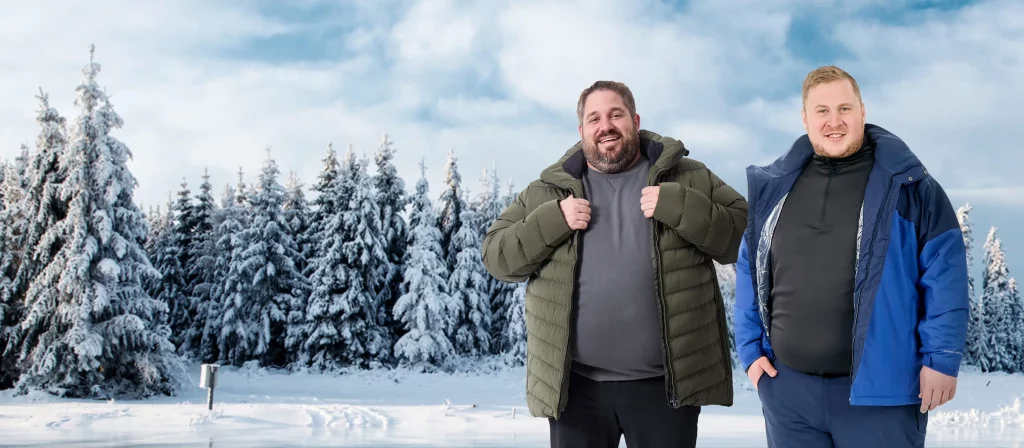 Zwei Männer mit Übergröße in warmen Winterjacken stehen vor einem verschneiten Wald. Der Boden ist mit Schnee bedeckt.