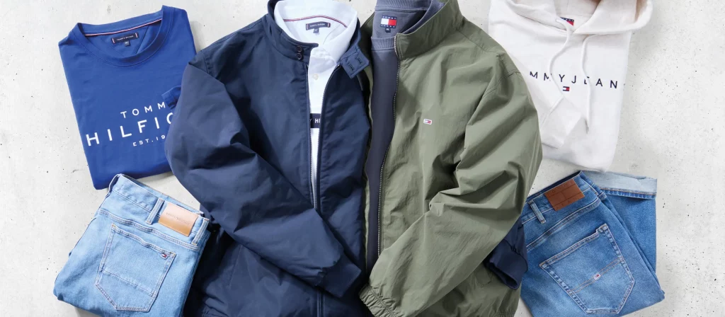 Gelegte Kleidungsstücke: Blaues T-Shirt, weißer Hoodie, zwei Jeans in unterschiedlicher Waschung, grüne und blaue Blousonjacken, weißes Polo-Shirt und dunkelblauer Pullover.