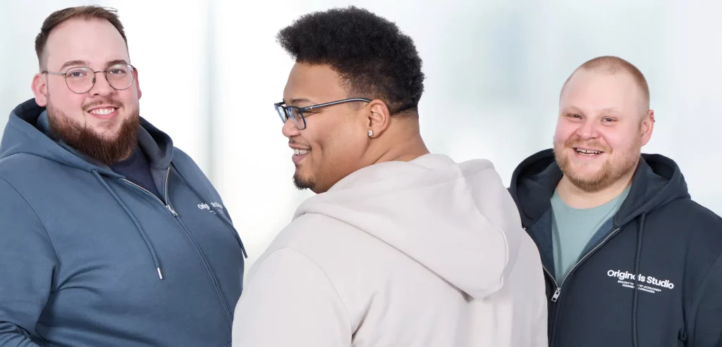 Drei Männer mit Übergröße in komfortablen Reise-Outfits: Links eine blaue Sweatjacke mit Jeans, in der Mitte ein Mann mit beiger Jogginghose und passender Sweatjacke, rechts eine schwarze Sweatjacke mit grünem T-Shirt und Jeans.