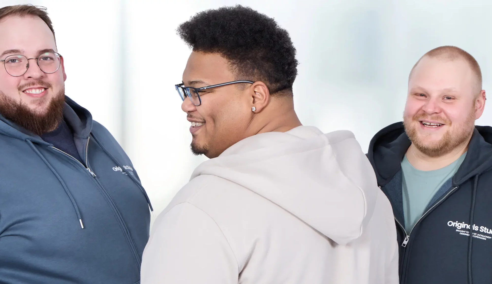 Drei Männer mit Übergröße in komfortablen Reise-Outfits: Links eine blaue Sweatjacke mit Jeans, in der Mitte ein Mann mit beiger Jogginghose und passender Sweatjacke, rechts eine schwarze Sweatjacke mit grünem T-Shirt und Jeans.