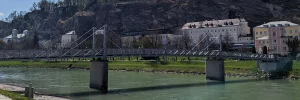 Blick auf die Salzach mit einer Fußgängerbrücke, im Hintergrund Hotels und Wohnhäuser in Salzburg.