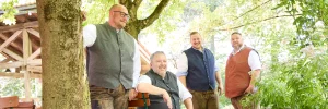 Vier Männer in traditioneller Tracht sitzen im Biergarten unter Kastanienbäumen und genießen das bayerische Lebensgefühl