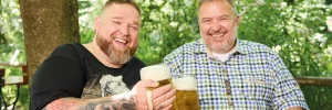 Zwei Männer in Lederhosen und Hemd/T-Shirt stoßen auf dem Volksfest mit einer Maß Bier an.
