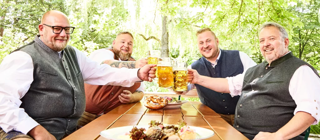 Vier Männer in Tracht stoßen im Biergarten mit Maß Bier an und genießen eine traditionelle Brotzeit.
