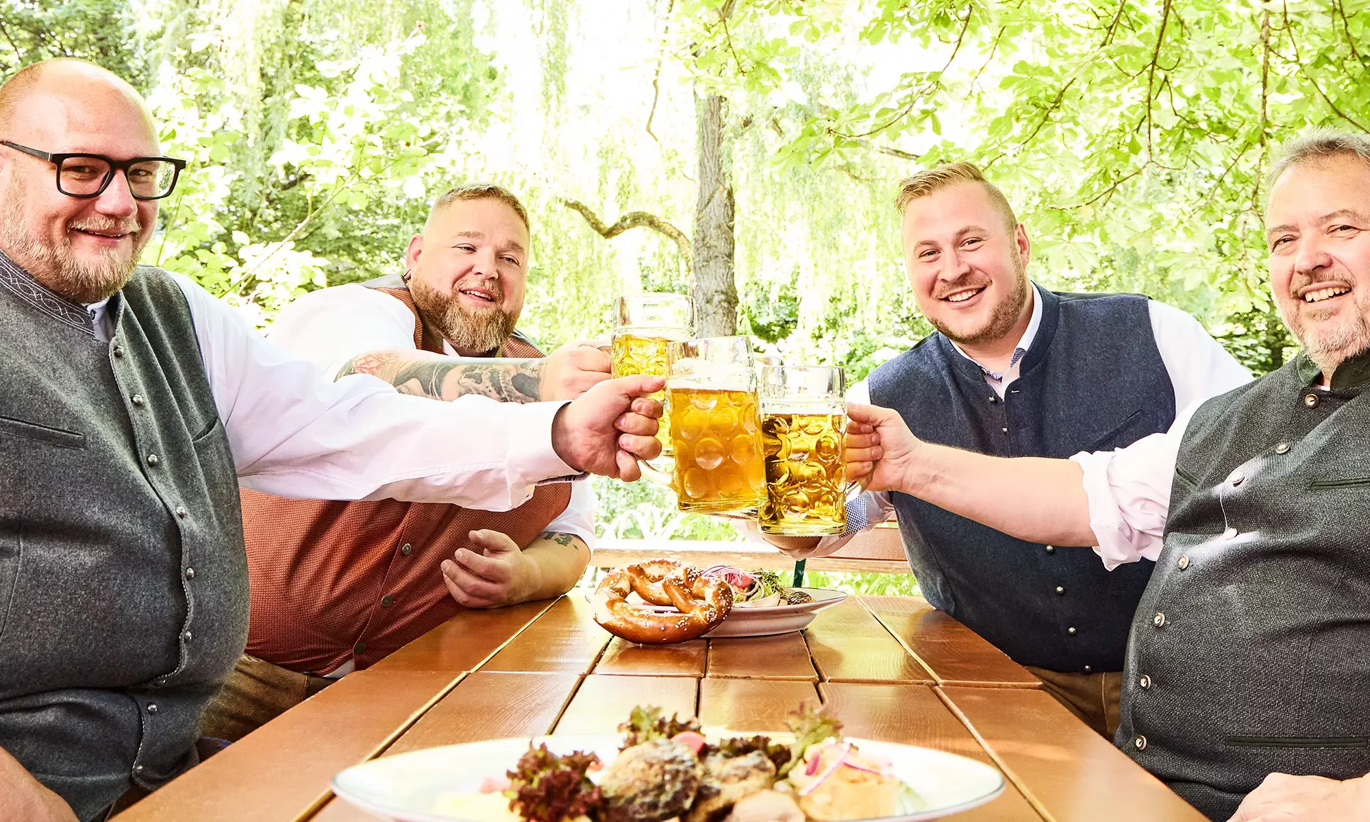 Vier Männer in Tracht stoßen im Biergarten mit Maß Bier an und genießen eine traditionelle Brotzeit.