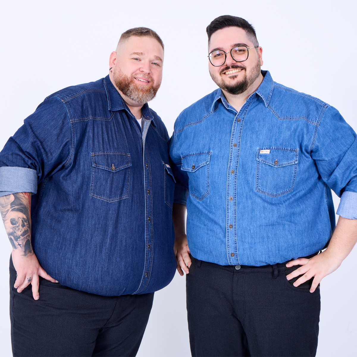 Zwei kräftige Männer stehen nebeneinander und lächeln freundlich in die Kamera. Beide tragen Jeanshemden – einer in Dunkelblau mit tätowiertem Unterarm, der andere in Hellblau mit Brille und Bart.
