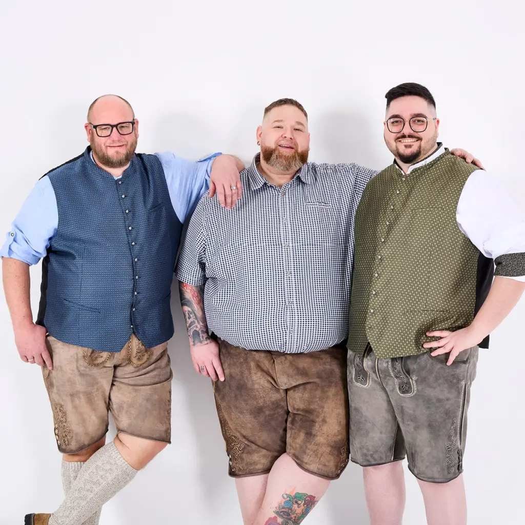 Drei gut gelaunte Männer in Trachtenoutfits – Lederhosen, Hemden und Westen – stehen entspannt nebeneinander und posieren vor hellem Hintergrund, ein lebendiger Moment voller Gemeinschaft und Stil