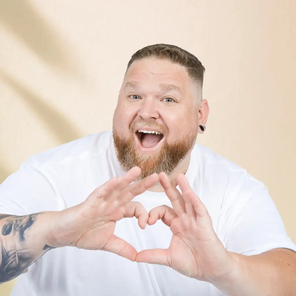 Mann mit Bart und weißem Shirt zeigt Herzgeste – authentisches Model für große Größen bei Pfundskerl.