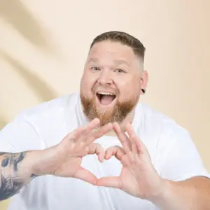 Mann mit Bart und weißem Shirt zeigt Herzgeste – authentisches Model für große Größen bei Pfundskerl.