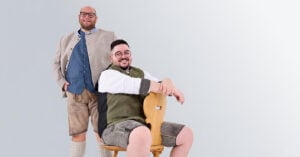 Zwei Männer in authentischer bayerischer Tracht posieren fröhlich: Ein Mann steht mit Janker und Lederhose, der andere sitzt auf einem Holzstuhl mit grüner Weste und kurzer Hirschlederhose