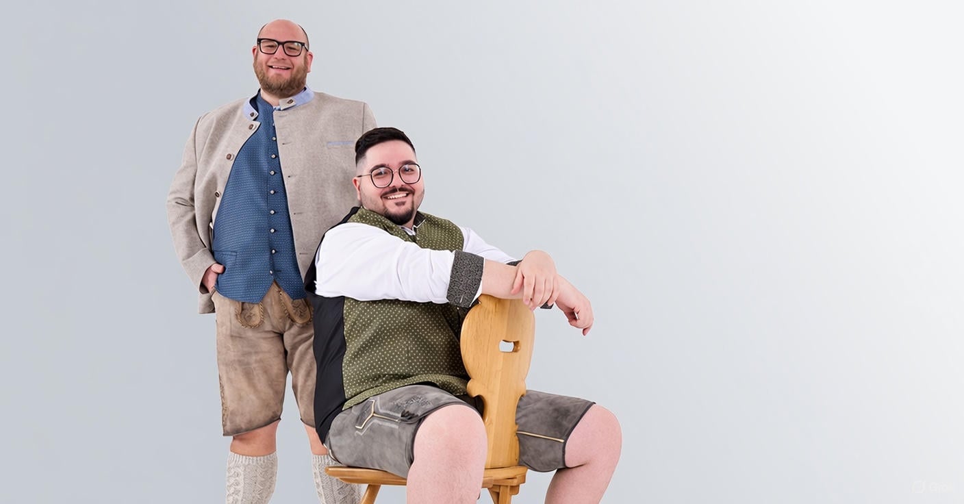 Zwei Männer in authentischer bayerischer Tracht posieren fröhlich: Ein Mann steht mit Janker und Lederhose, der andere sitzt auf einem Holzstuhl mit grüner Weste und kurzer Hirschlederhose