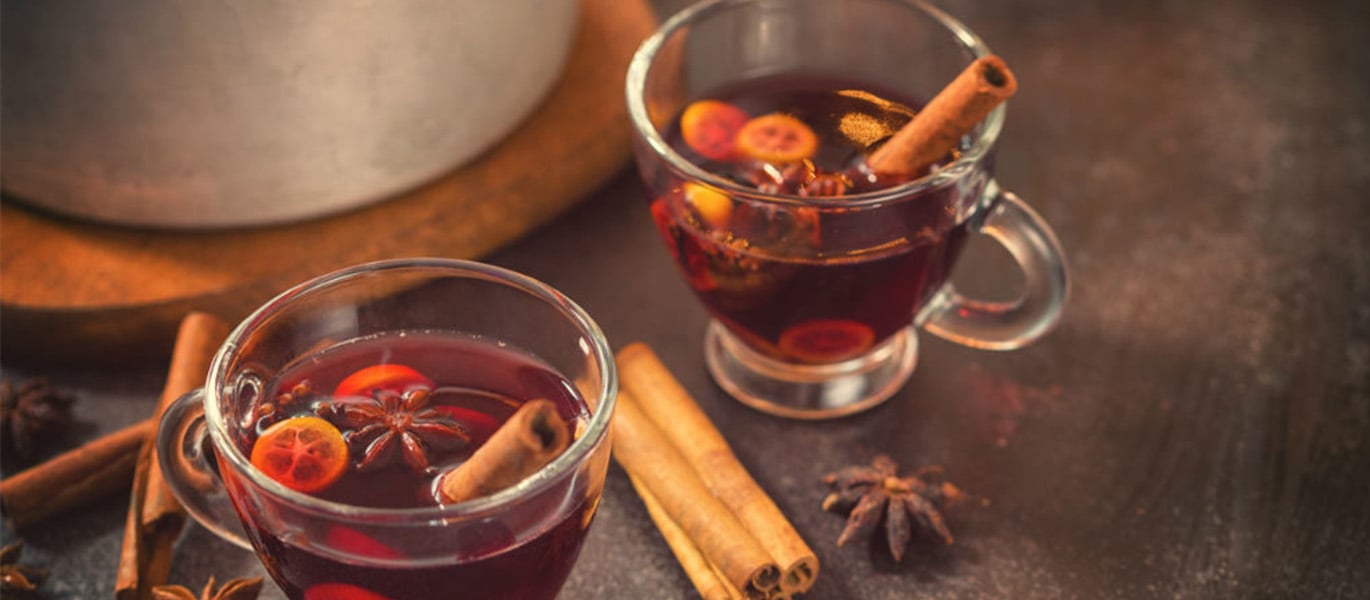 Zwei Tassen heißer Glühwein mit Zimtstangen, Sternanis und Orangenscheiben auf einem dunklen Tisch.