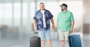 Zwei Männer mit Format stehen lachend mit Rollkoffern in einer hellen Flughafenhalle, einer im gemusterten Kurzarmhemd, einer im mintgrünen Poloshirt