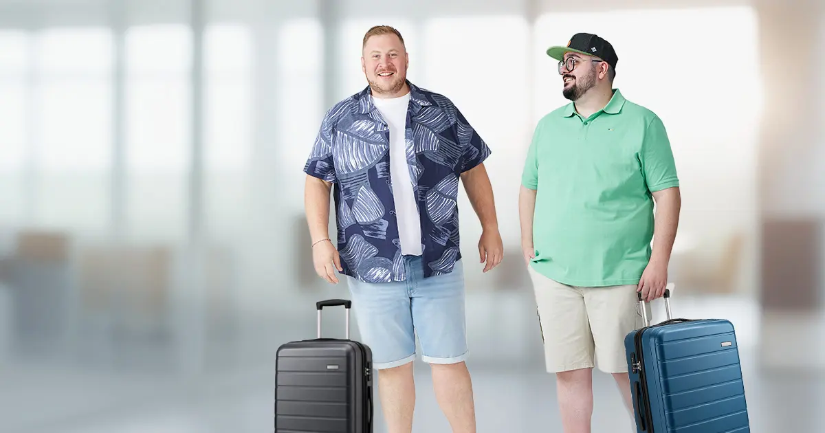 Zwei Männer mit Format stehen lachend mit Rollkoffern in einer hellen Flughafenhalle, einer im gemusterten Kurzarmhemd, einer im mintgrünen Poloshirt
