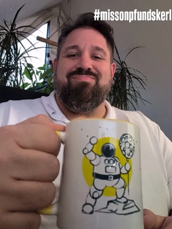 Mann mit Bart und weißem Shirt hebt seine Pfundskerl-Sammeltasse mit Astronaut und Mondballon