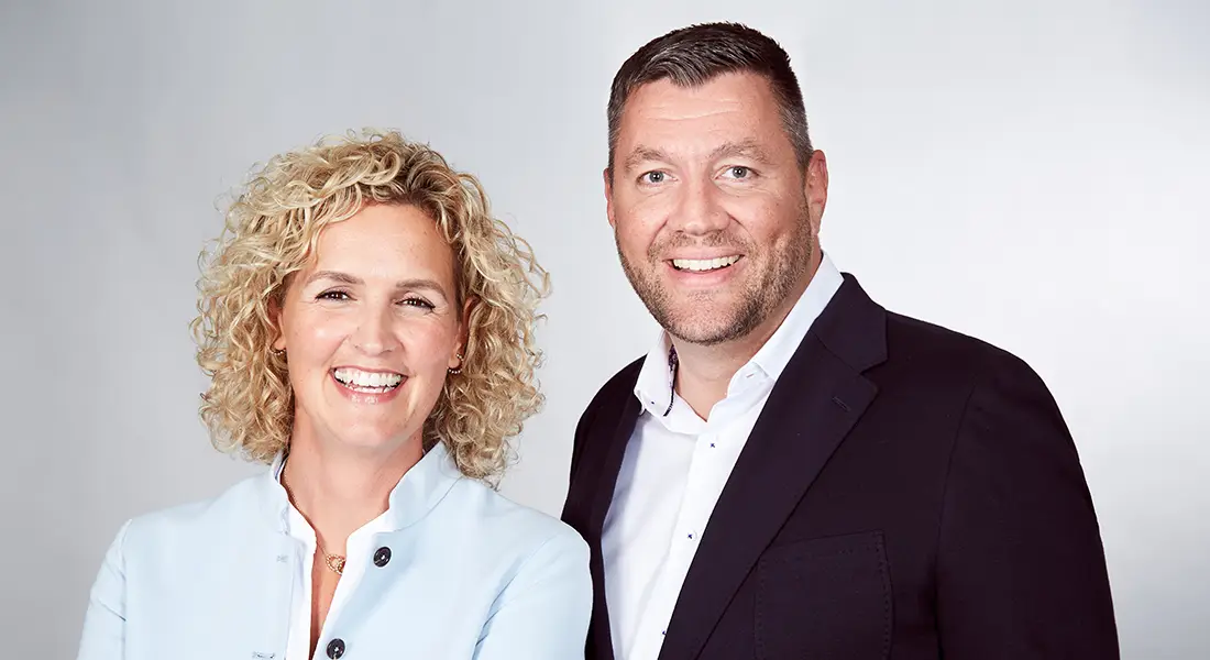 Eine Frau und ein Mann in eleganter Businesskleidung stehen lächelnd gemeinsam vor einem hellen Hintergrund.