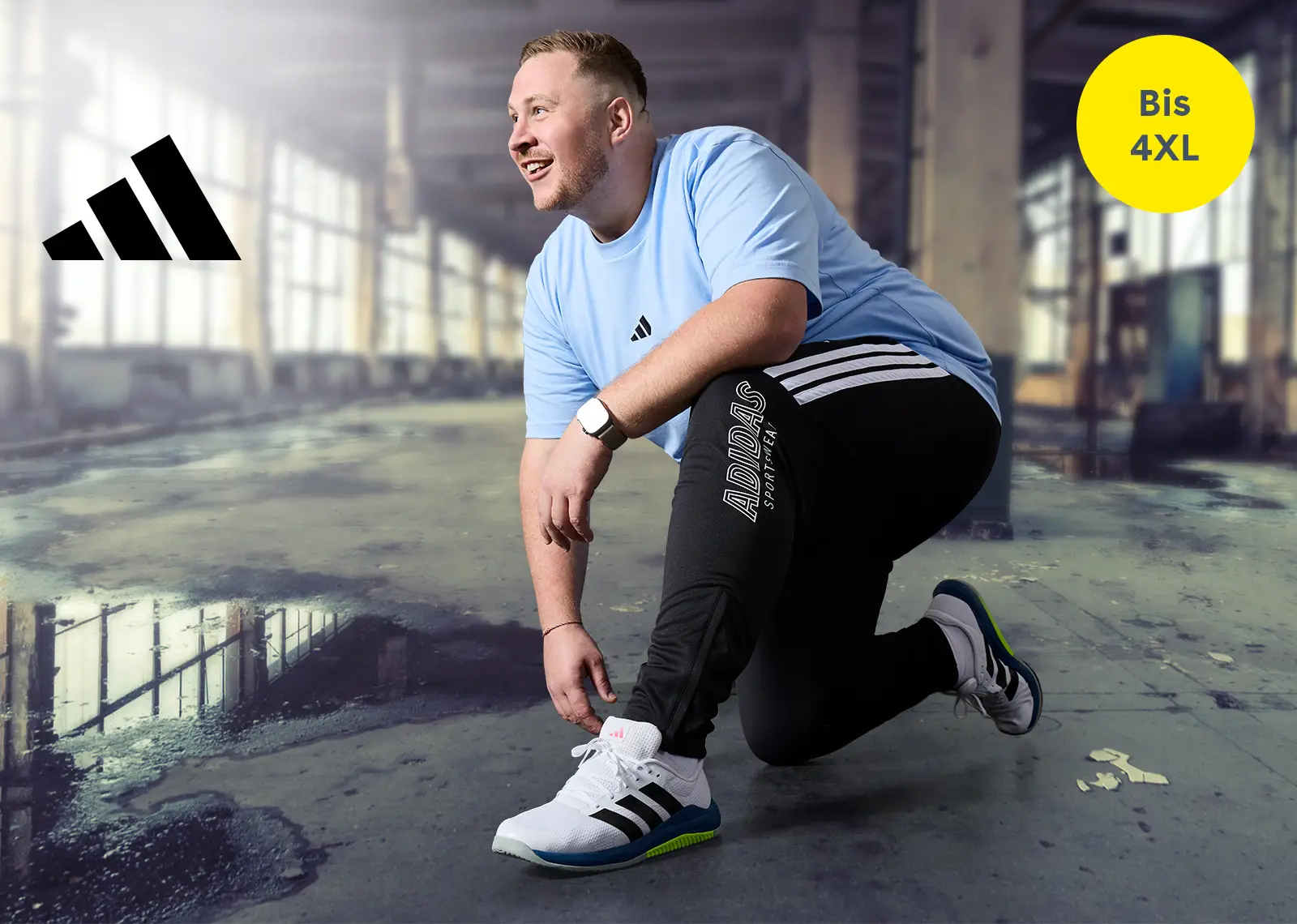 Mann in großer Größe kniet im Fitnessstudio und trägt adidas Trainingsanzug und Sneaker – Sportbekleidung bis 4XL