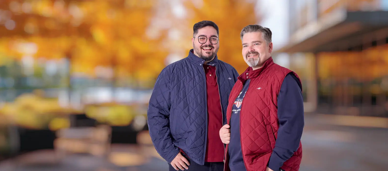 Zwei Männer in herbstlicher Herrenmode mit blauer Steppjacke und roter Steppweste vor herbstlicher Kulisse.