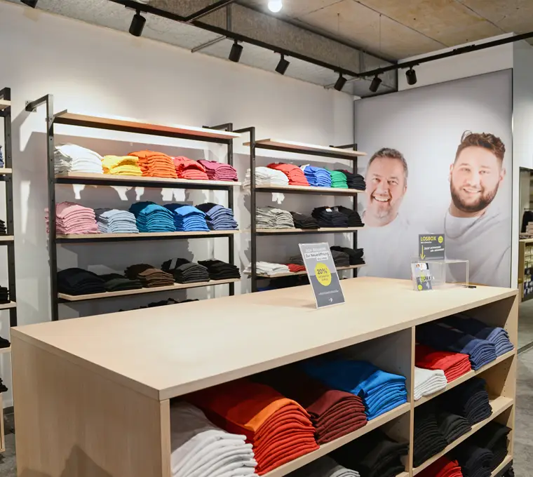 Verkaufsfläche mit farblich sortierten T-Shirts in großen Größen – im Hintergrund lächelndes Pfundskerl-Team
