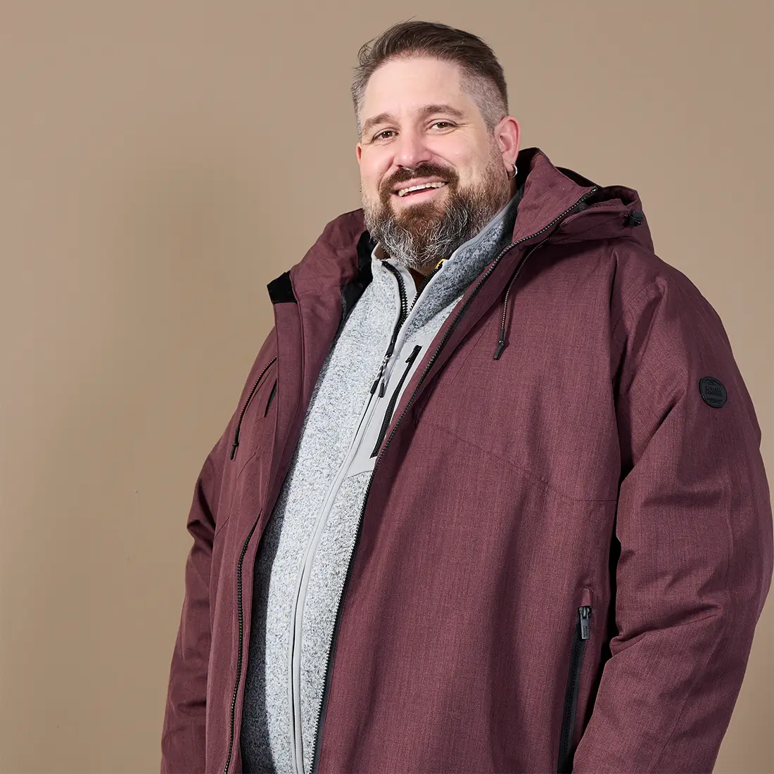 Mann in großer Größe trägt eine bordeauxrote Winterjacke mit Kapuze über einer hellgrauen Fleecejacke.