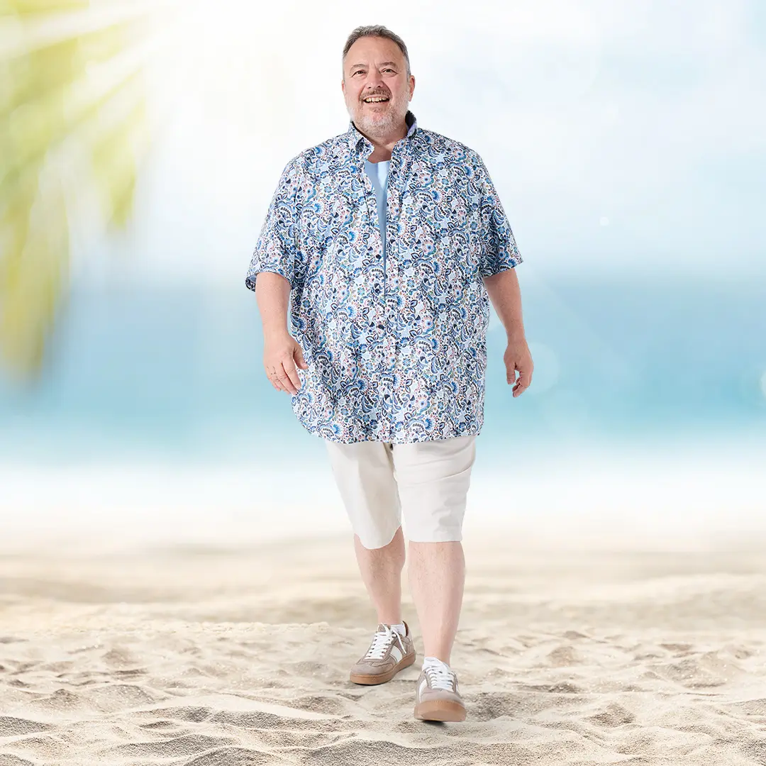 Ein lächelnder Mann in blau-weiß gemusterter Kurzarmbluse und hellen Bermudas genießt den Strand – stilvoller Urlaubslook ab 3XL.