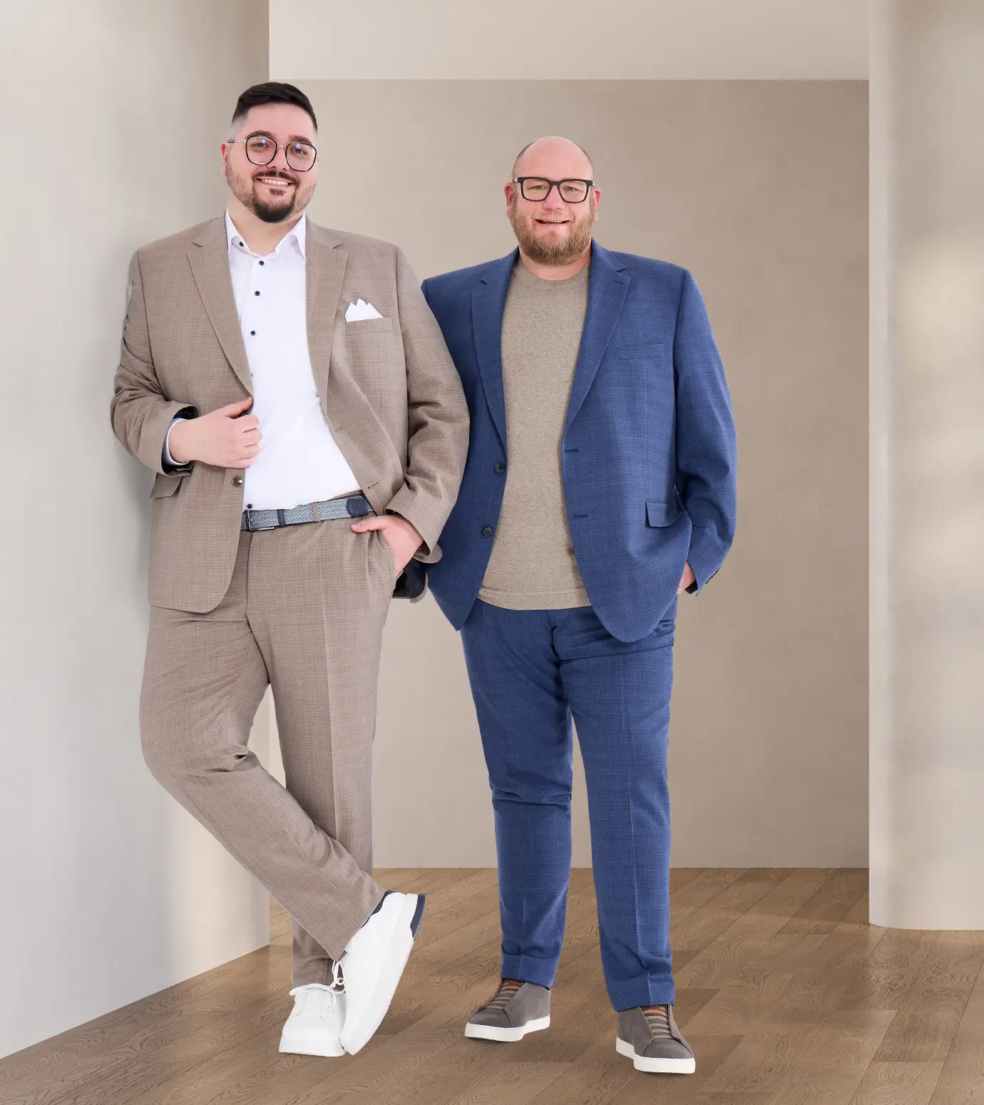 Zwei Männer in stilvollen Anzügen, einer in Beige mit weißen Sneakern und der andere in Blau mit grauen Schuhen, posieren vor einer minimalistischen Wand.