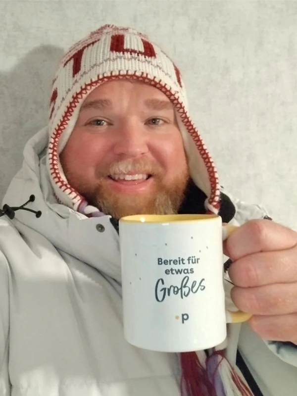 Lächelnder Mann im Winteroutfit hält den Pfundskerl-Sammeltassenbecher mit der Aufschrift „Bereit für etwas Großes“ in die Kamera