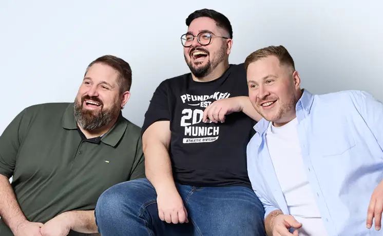 Drei gut gelaunte Männer in großen Größen sitzen gemeinsam vor hellem Hintergrund und tragen lässige Pfundskerl-Outfits