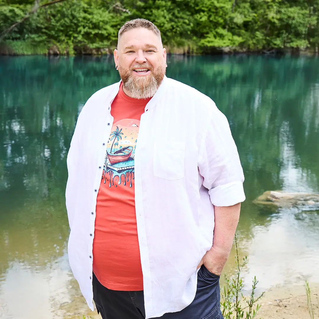 Mann in rotem Printshirt und weißem Leinenhemd am Wasser – sommerlicher Look mit lockerem Fit für große Größen.