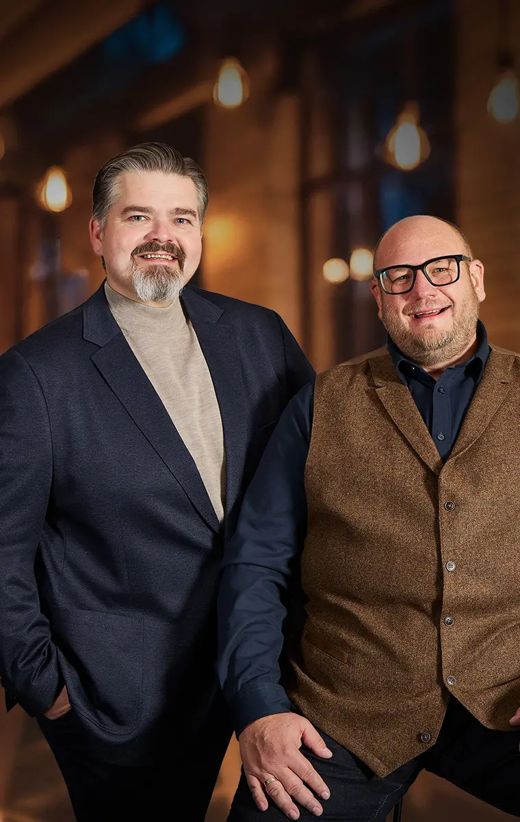 Zwei Männer in großen Größen in eleganten Weihnachtsoutfits vor warmem Lichterhintergrund