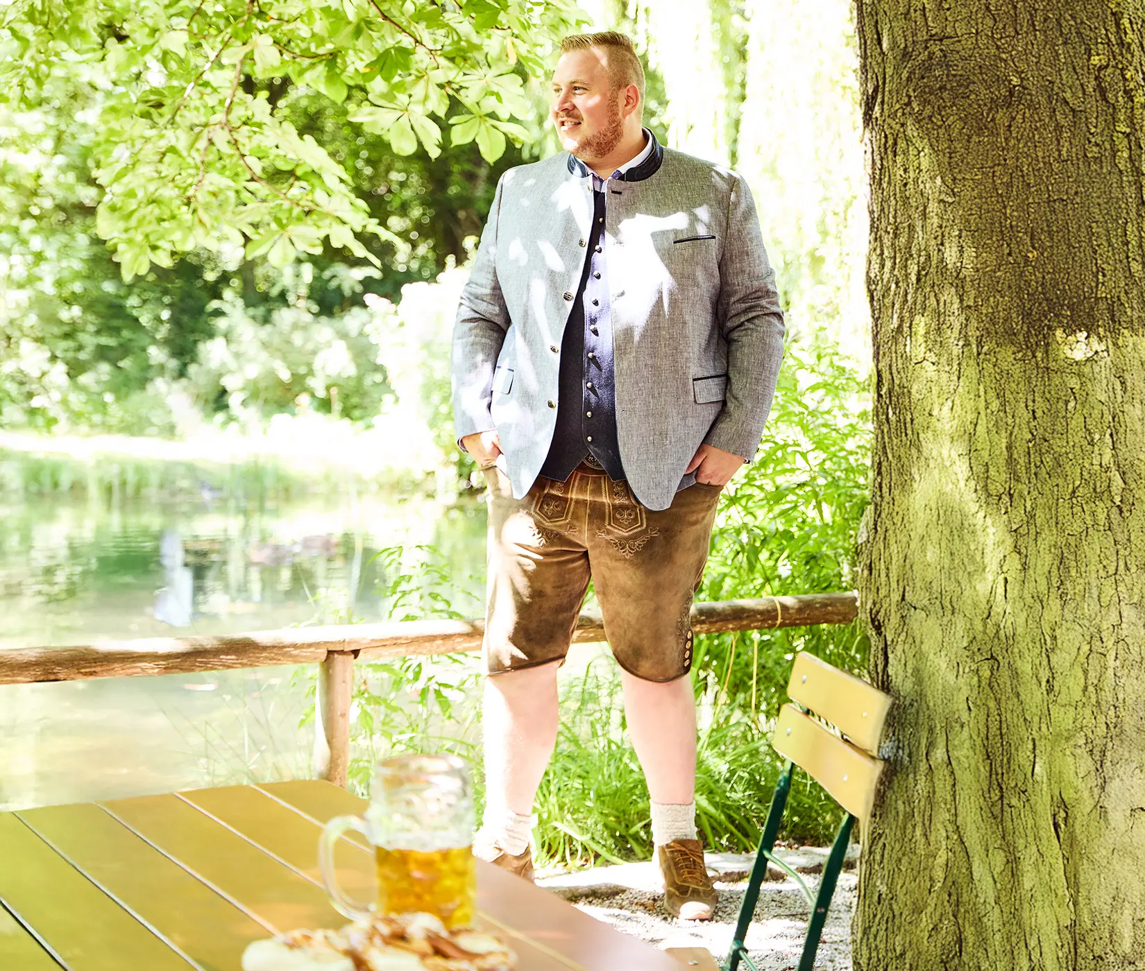 Mann mit hellem Trachtenjanker, dunkelblauem Hemd und traditioneller Lederhose steht am See – stilvoller Look für kräftige Herren.