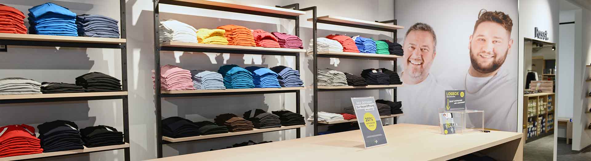 T-Shirt-Regale im Pfundskerl Store mit bunter Größenauswahl und freundlichem Team-Foto im Hintergrund