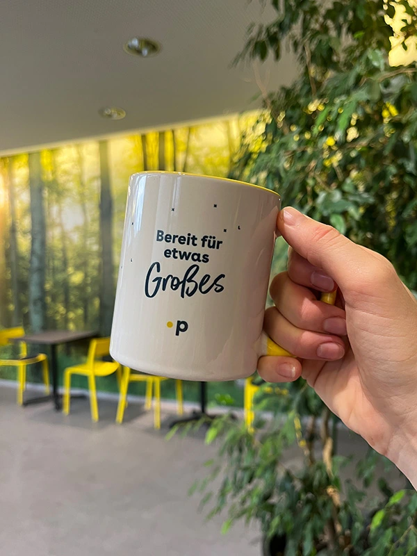 Hand hält Pfundskerl-Sammeltasse mit der Aufschrift „Bereit für etwas Großes“ vor einem grünen Hintergrund