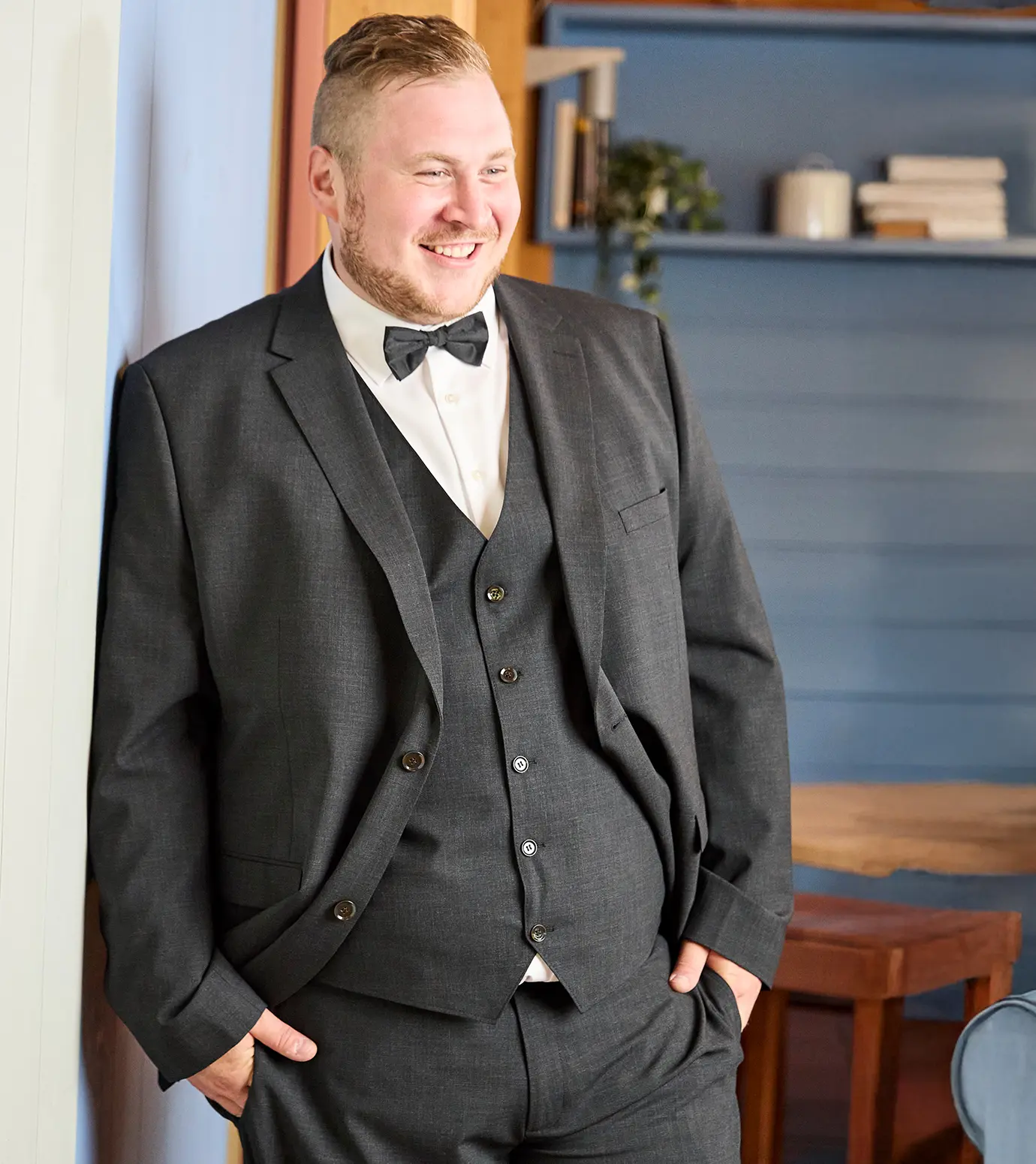 Person in dunkelgrauem Anzug mit weißem Hemd und Fliege lehnt an Wand, im Hintergrund Bücherregal mit Dekoration.