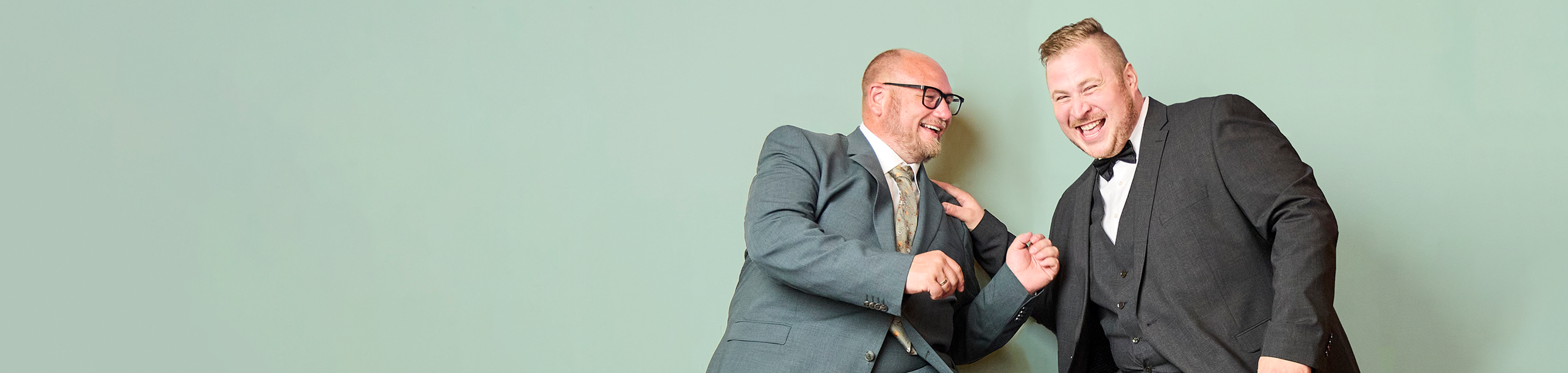 Zwei lachende Männer in eleganten Anzügen, einer in Grün mit gemusterter Krawatte, der andere in Dunkelgrau mit Fliege, gestikulieren vor grünem Hintergrund.