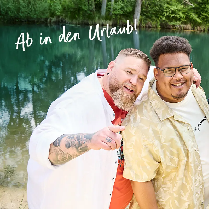 Zwei lächelnde Männer in Sommermode posieren am See mit Schriftzug „Ab in den Urlaub!“ – mobile Header-Version mit XXL-Charme.