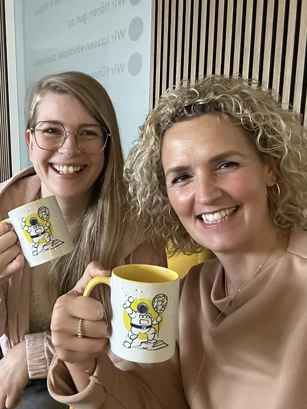 Zwei lachende Frauen halten ihre Pfundskerl-Sammeltassen mit Astronautenmotiv in die Kamera