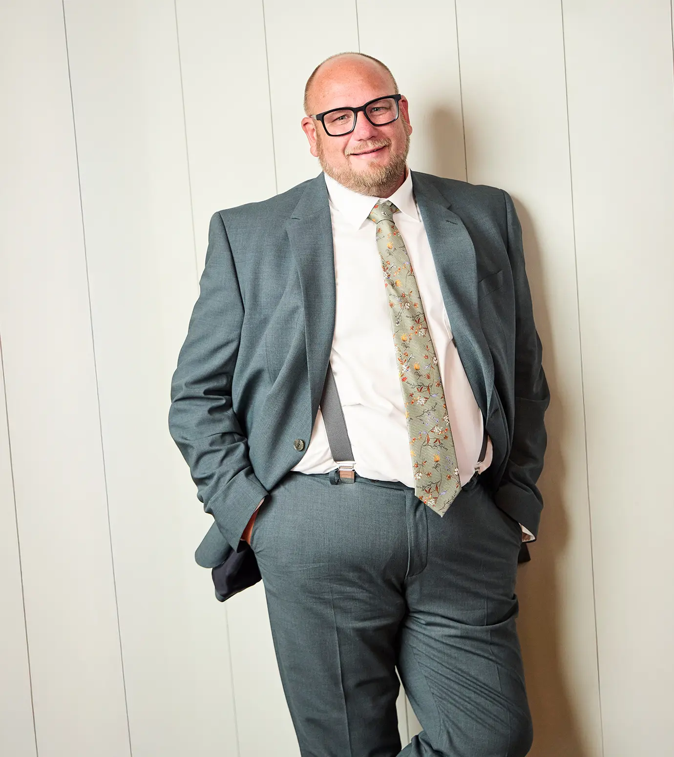 Person im grünen Anzug mit floraler Krawatte und Hosenträgern lehnt an hell gestrichener Holzverkleidung.