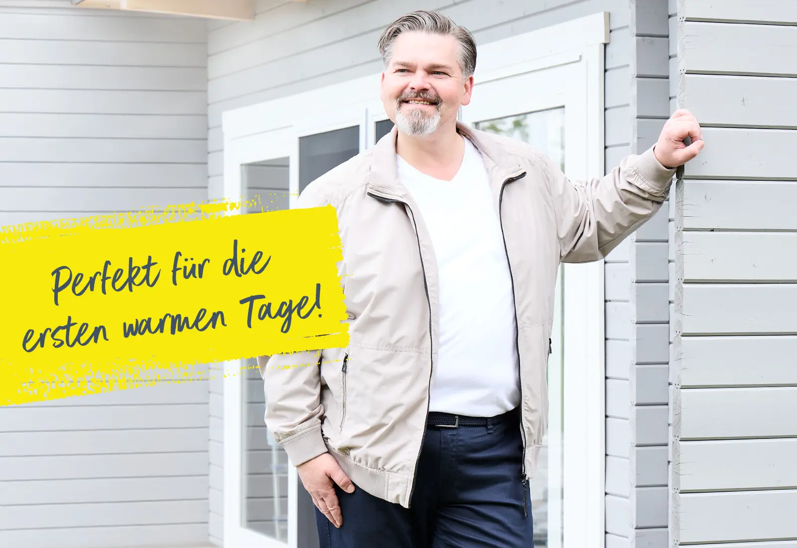 Mann in beiger Übergangsjacke lehnt lächelnd an Hauswand