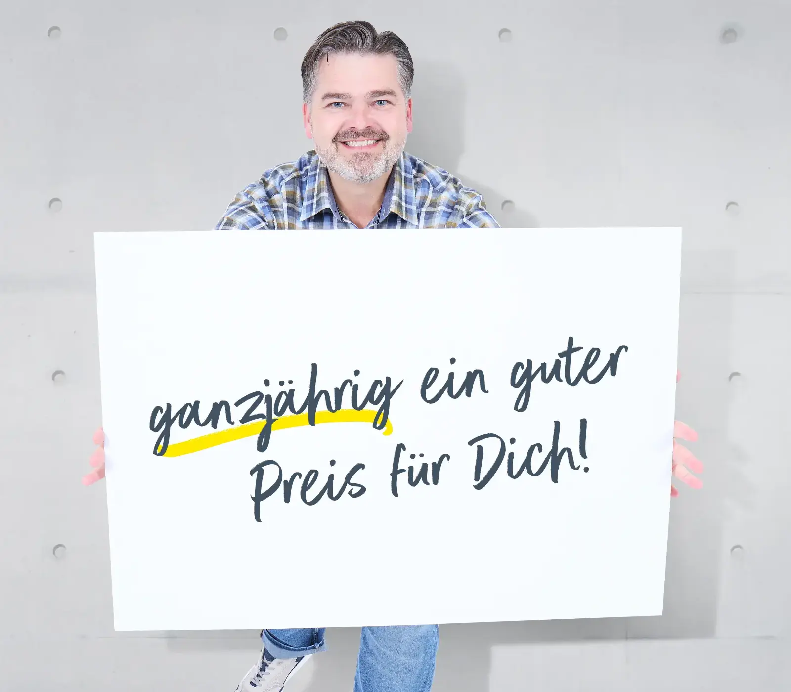Mann in blauem Karohemd hält Schild mit Aufschrift: ganzjährig ein guter Preis für Dich!, vor heller Wand.