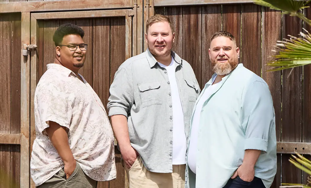 Drei Männer in großen Größen tragen luftige Sommerhemden und entspannte Outfits vor Holztor im Freien