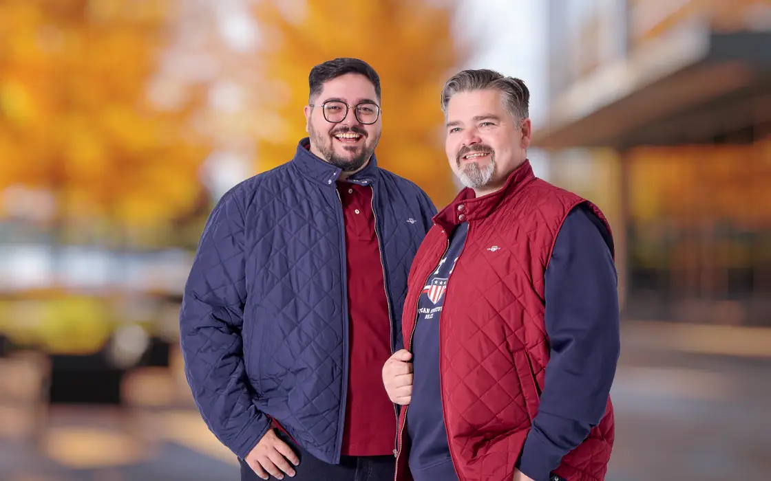 Zwei Männer in herbstlicher Herrenmode mit blauer Steppjacke und roter Steppweste vor herbstlicher Kulisse.